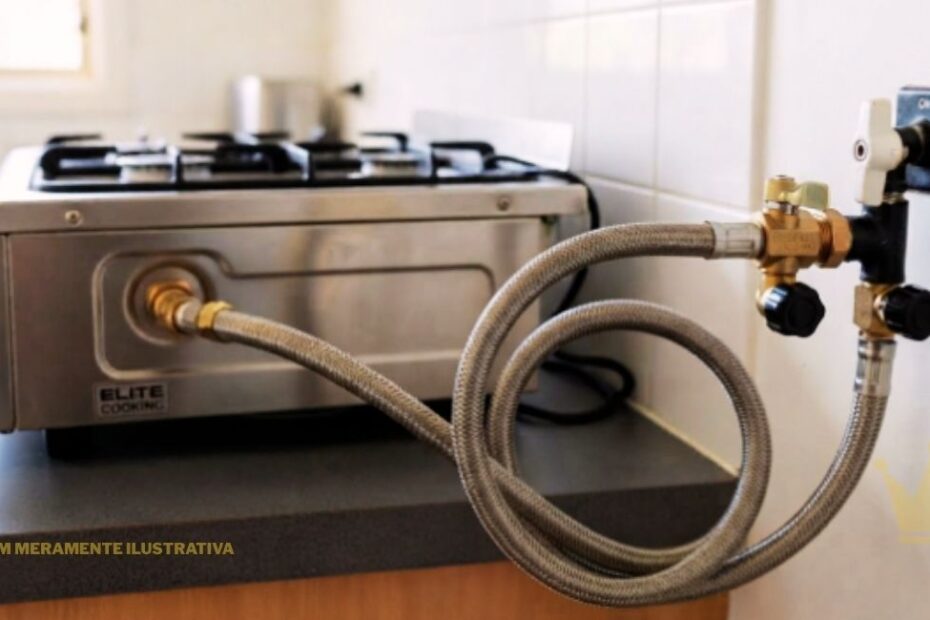 "Vista aproximada de uma mangueira de gás flexível de malha de aço instalada em uma cozinha residencial. A mangueira metálica trançada faz uma curva e conecta a entrada traseira de um fogão cooktop de bancada em aço inox a um registro e válvula de segurança fixados na parede de azulejos claros."